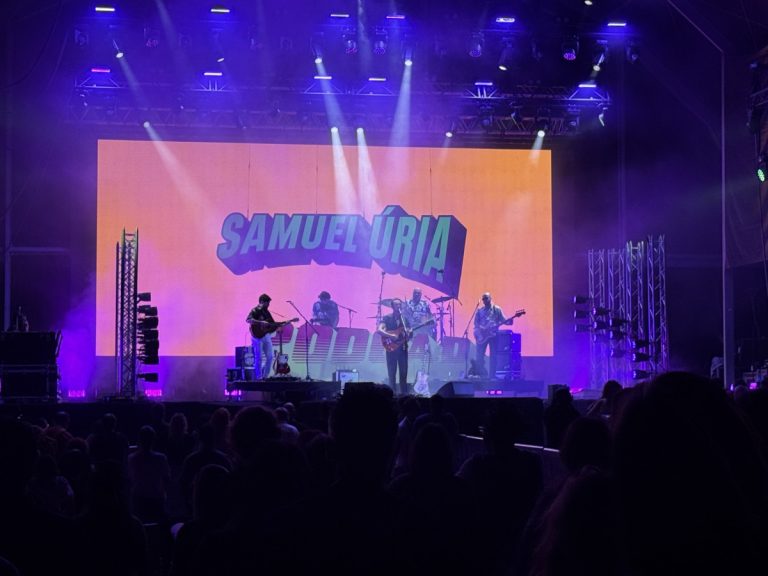 Concert stage with a band performing under purple lights; large orange screen behind reads 'SAMUEL URIA' in blue letters; audience silhouettes in foreground.
