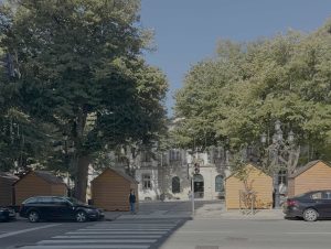 Tree-lined city street with wooden market stalls, parked cars, and a crosswalk in front of a pale building.
