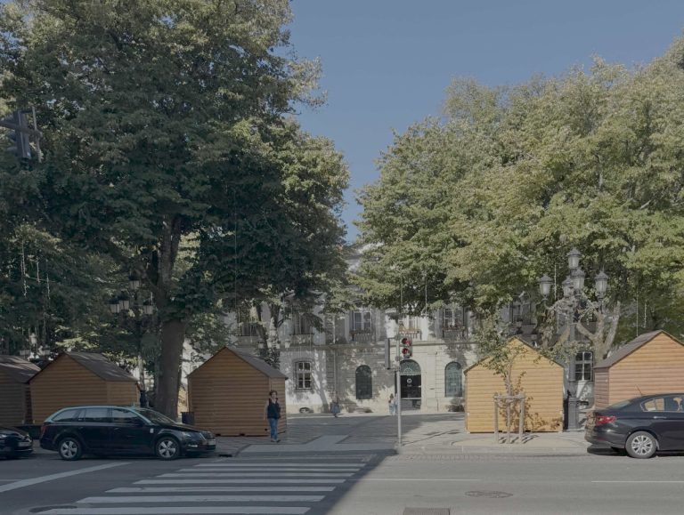 Tree-lined city street with wooden market stalls, parked cars, and a crosswalk in front of a pale building.