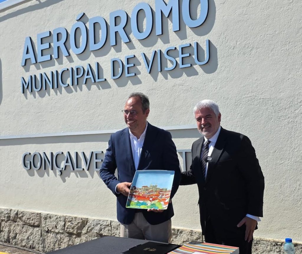 Outside Aeródromo Municipal de Viseu, two men in suits exchange a colorful decorative plate and smile.