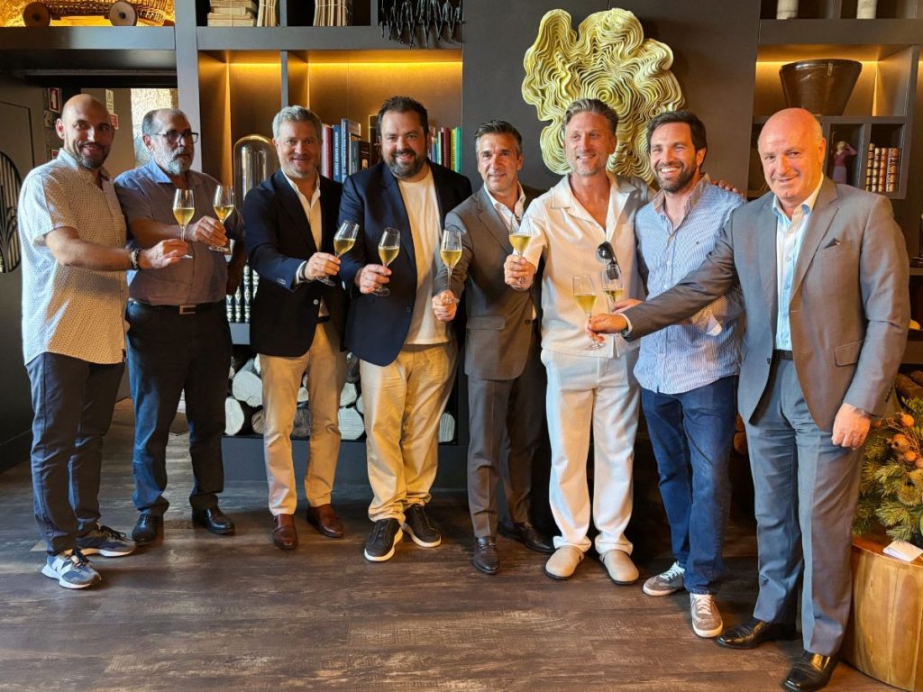 Eight men in smart casual attire raise champagne flutes in a toast in a stylish lounge with shelves and art behind them.
