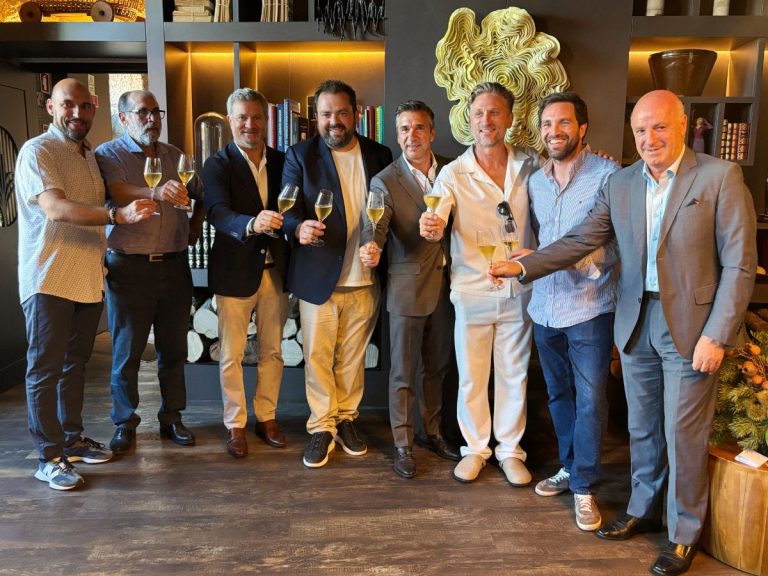 Eight men in smart casual attire raise champagne flutes in a toast in a stylish lounge with shelves and art behind them.