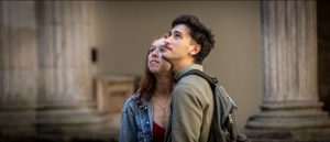 Young couple near classical stone columns on a campus; the man with a backpack gazes upward as the woman looks on.