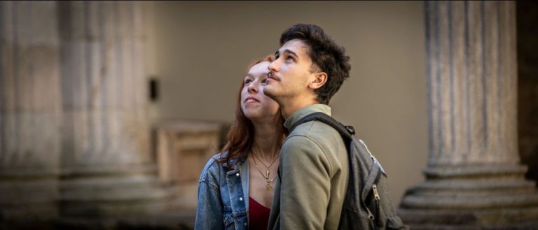 Young couple near classical stone columns on a campus; the man with a backpack gazes upward as the woman looks on.