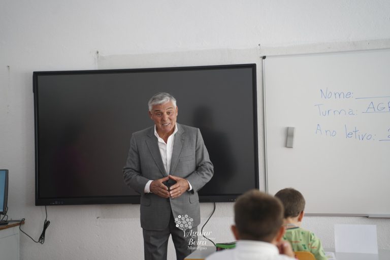 Male teacher in a gray suit presenting to students in a classroom with a large wall-mounted screen behind him.