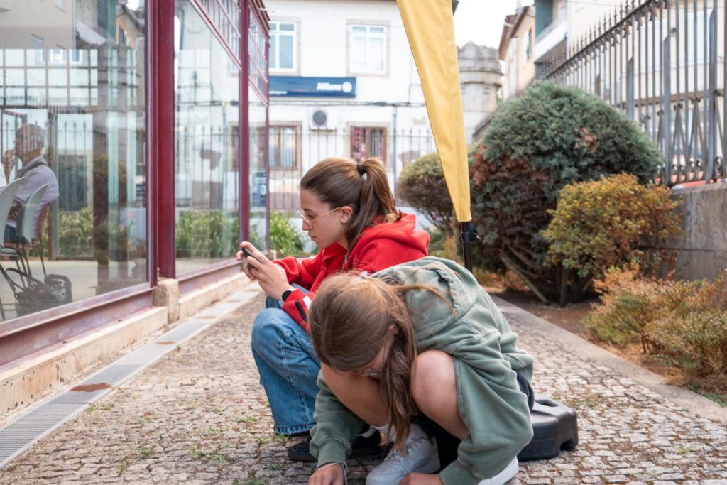 Two girls on a cobblestone sidewalk beside a glass storefront; one in a red hoodie uses a phone while the other sketches on the ground with chalk.