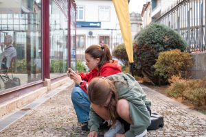 Two girls on a cobblestone sidewalk beside a glass storefront; one in a red hoodie uses a phone while the other sketches on the ground with chalk.