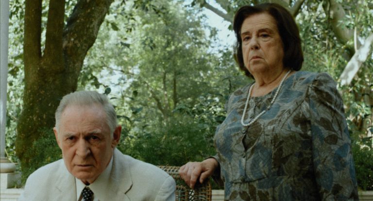 Elderly man seated in a light suit with a serious expression, while an older woman in a patterned dress with a pearl necklace stands beside him in a lush garden.