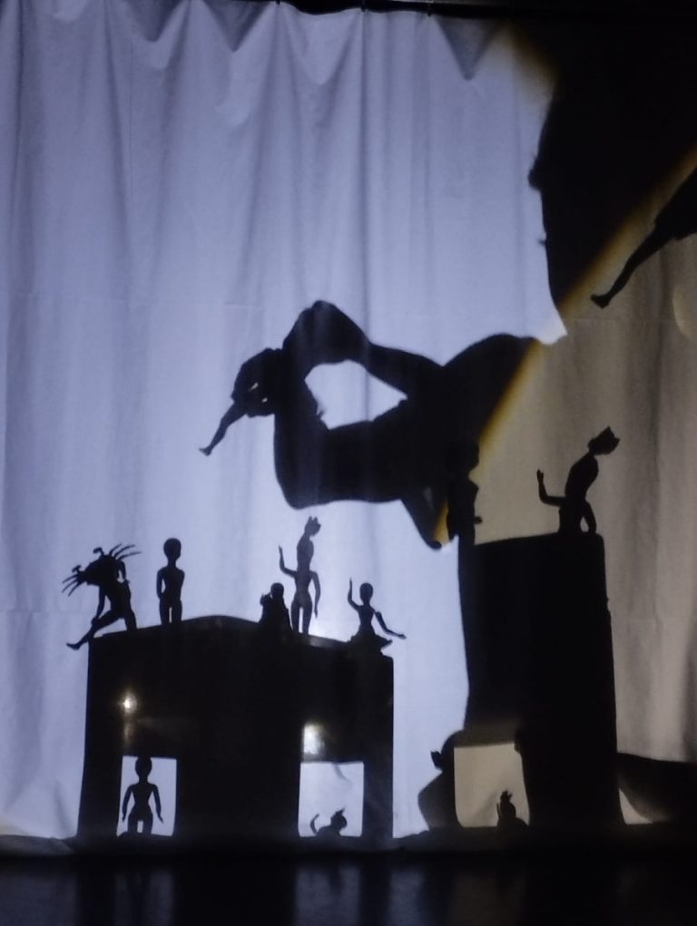 Silhouettes of performers and figures on a stage, with a large figure leaping overhead against a light screen and smaller characters on platforms below.