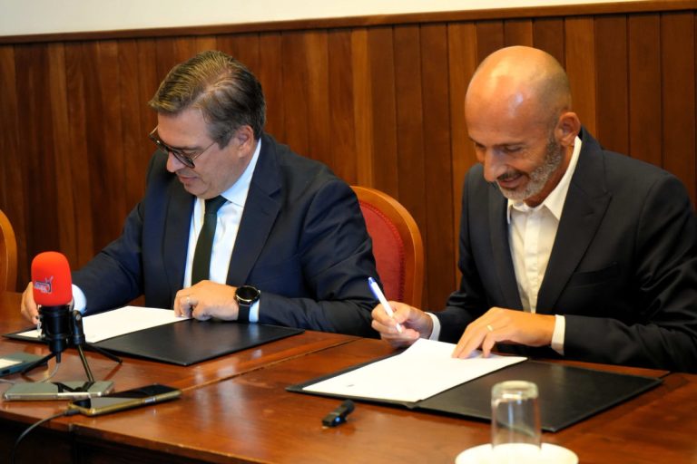 Two men in dark suits sign documents at a wooden table, with microphones and recording devices nearby.