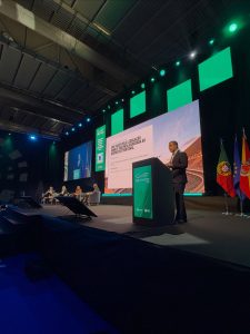 Speaker at a podium addressing a conference on stage with a large screen, panelists, and flags in the background