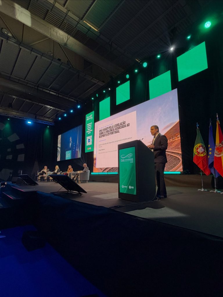 Speaker at a podium addressing a conference on stage with a large screen, panelists, and flags in the background