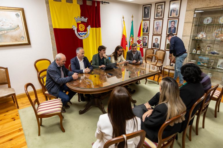 Seven people sit around a polished conference table in a formal room, with flags on the wall and framed portraits nearby.