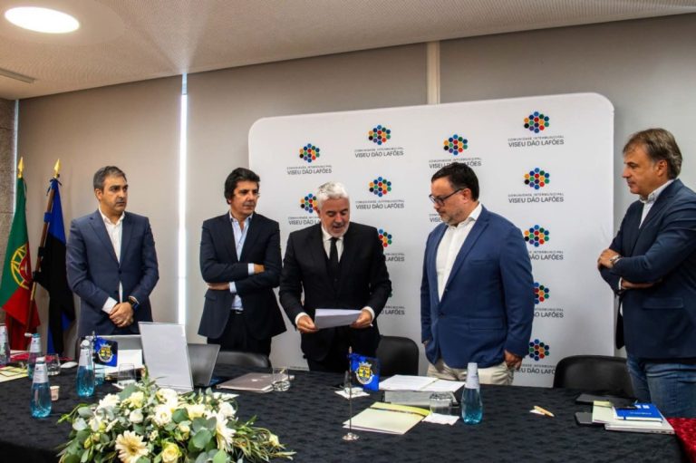 Five men in suits stand around a conference table with documents and laptops, in front of a branded backdrop with logos and flags nearby.