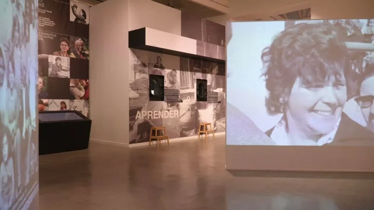 Museum gallery with a large video projection of a smiling person on the right and collage walls with photos and text like 'APRENDER'. Two wooden stools sit nearby.