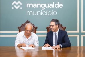 Two men sit at a wooden table signing documents in front of a teal wall with a 'Mangualde Município' sign.