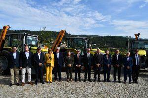 Group of professionals in formal attire standing in a row on a stone lot with yellow tractors behind them and a rural backdrop of hills and clear sky.