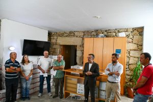 Eight people stand in a stone-walled room listening to a man in a suit who is speaking near a wooden cabinet and shelves.