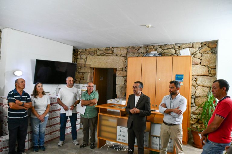 Eight people stand in a stone-walled room listening to a man in a suit who is speaking near a wooden cabinet and shelves.