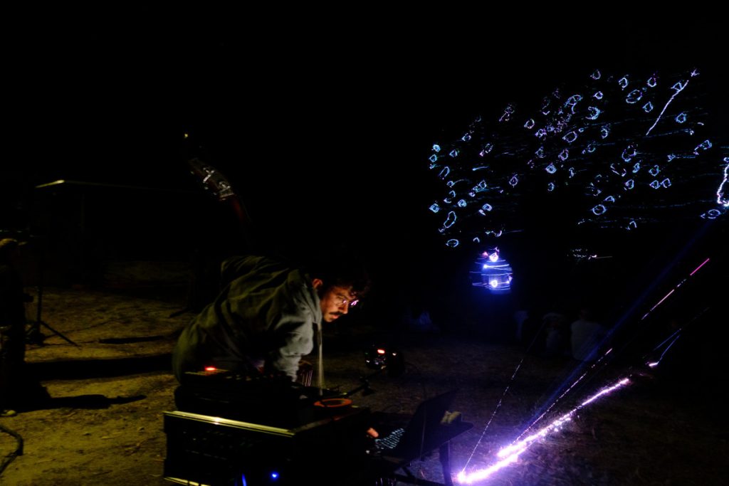 DJ wearing a gray hoodie leans over mixing gear at night, with blue neon tree lights in the background.