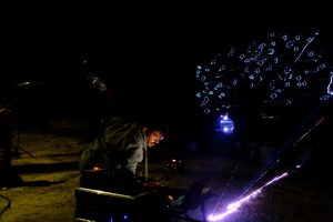 DJ wearing a gray hoodie leans over mixing gear at night, with blue neon tree lights in the background.