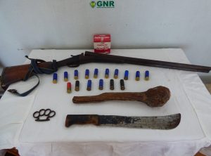 Seized weapons and ammunition displayed on a table under a GNR banner: a long rifle, a large curved blade, a rusted machete, knuckles, and multiple shotgun shells.