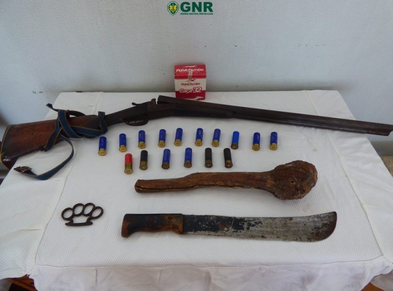 Seized weapons and ammunition displayed on a table under a GNR banner: a long rifle, a large curved blade, a rusted machete, knuckles, and multiple shotgun shells.