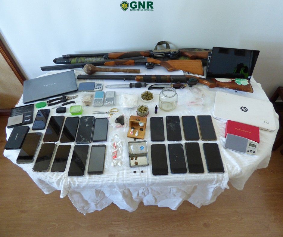 Table display of seized items: rifles, drugs in bags, jars, scales, and many smartphones with a laptop nearby and a GNR logo above.