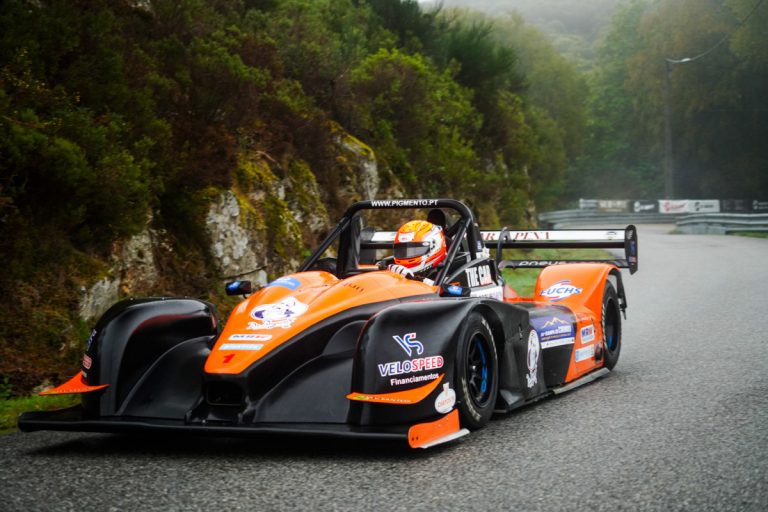 Orange and black open-cockpit race car speeding on a winding road, driver in orange helmet, sponsor decals visible on the bodywork.