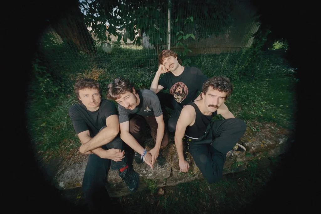 Four men with mustaches sit on a stone wall outdoors, posing for a group photo with greenery and a fence behind them.