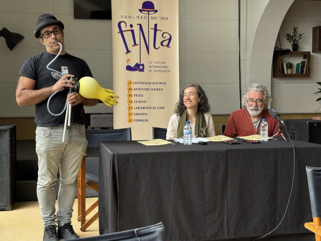 Man wearing a hat and glasses holds a device with a large yellow glove while two panelists sit at a table with water bottles and a fiNta banner in the background.