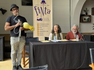 Man wearing a hat and glasses holds a device with a large yellow glove while two panelists sit at a table with water bottles and a fiNta banner in the background.
