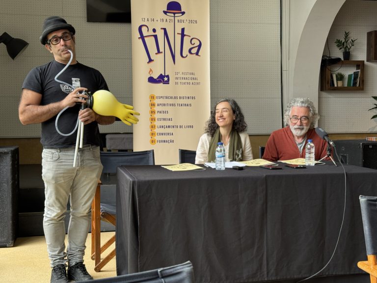 Man wearing a hat and glasses holds a device with a large yellow glove while two panelists sit at a table with water bottles and a fiNta banner in the background.