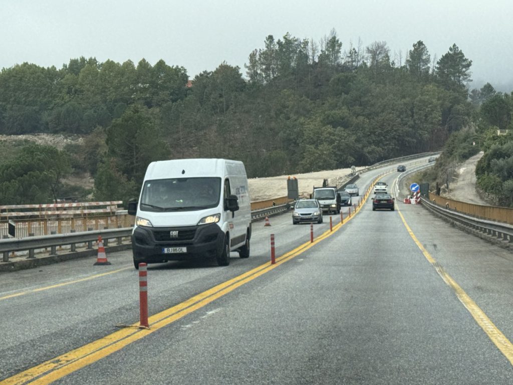 White van driving on a two-lane road under construction, orange traffic cones along the edge, with a forested hillside in the background.