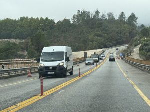 White van driving on a two-lane road under construction, orange traffic cones along the edge, with a forested hillside in the background.