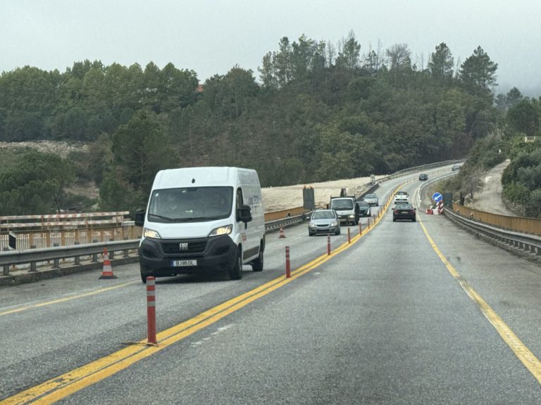 White van driving on a two-lane road under construction, orange traffic cones along the edge, with a forested hillside in the background.