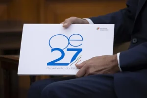 Person in a dark suit holds a white poster with the blue 'OE 27' logo and the text 'Orçamento do Estado'.