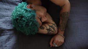 Person with teal curly hair lying on a dark quilt, arms tattooed and resting beside them on the bedbedspread.