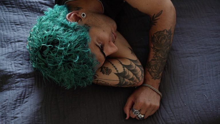 Person with teal curly hair lying on a dark quilt, arms tattooed and resting beside them on the bedbedspread.