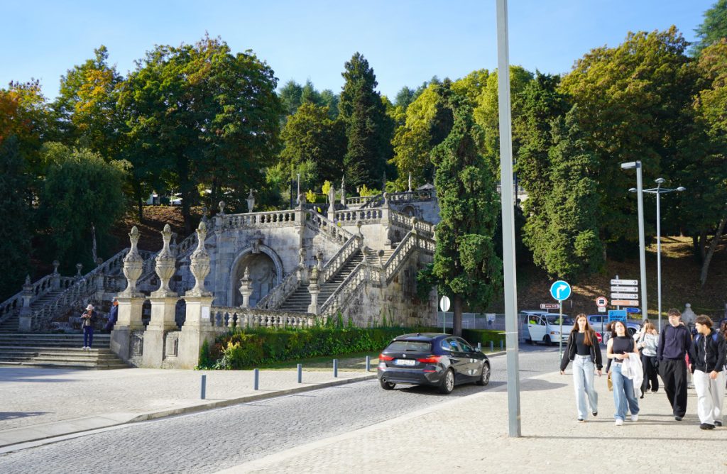 Stone staircase with ornate balustrades and a carved arch, set among green trees; pedestrians walk on the right.