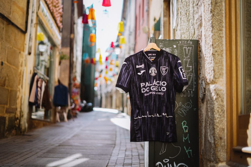 Black striped soccer jersey with 'PALÁCIO DO GELO' logo hanging on a hanger against a graffiti-covered utility box in a sunlit narrow alley with colorful banners overhead.