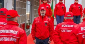 Bearded firefighter in a red uniform stands at the center among colleagues in red jackets during a ceremony.