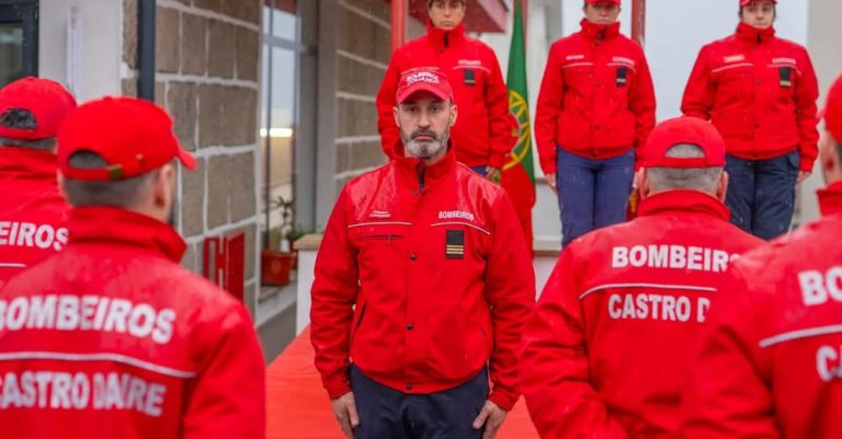 Bearded firefighter in a red uniform stands at the center among colleagues in red jackets during a ceremony.