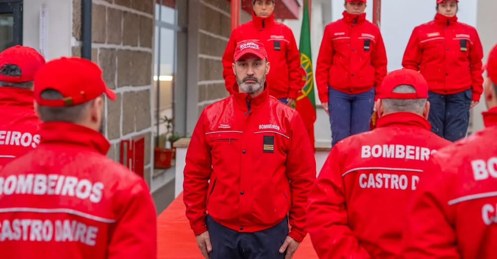 Bearded firefighter in a red uniform stands at the center among colleagues in red jackets during a ceremony.