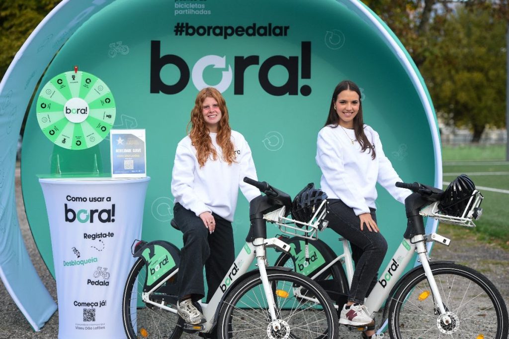 Two smiling women in white sweatshirts sit on white_bora bikes at a turquoise promotional booth with a green prize wheel behind them.