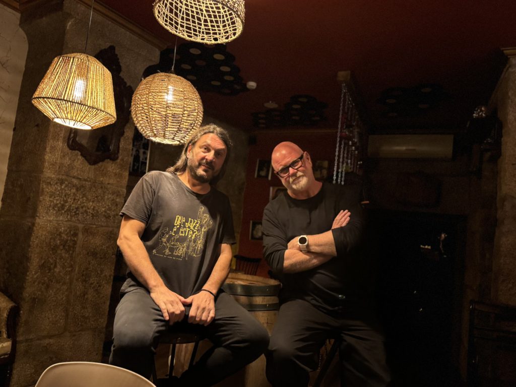 Two men sit on stools in a dim, rustic bar with wicker pendant lights overhead.