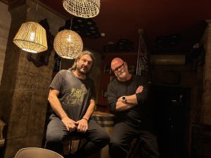 Two men sit on stools in a dim, rustic bar with wicker pendant lights overhead.