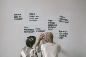 Two people stand with their backs to the camera, looking at white posters with Portuguese questions about dance on a wall.