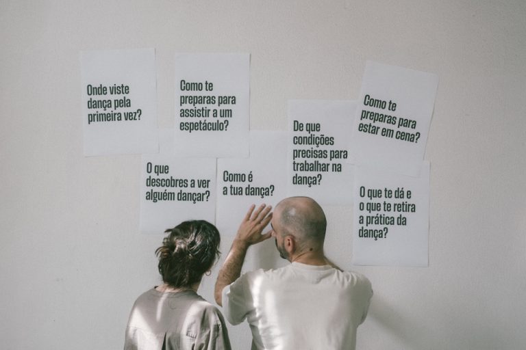 Two people stand with their backs to the camera, looking at white posters with Portuguese questions about dance on a wall.