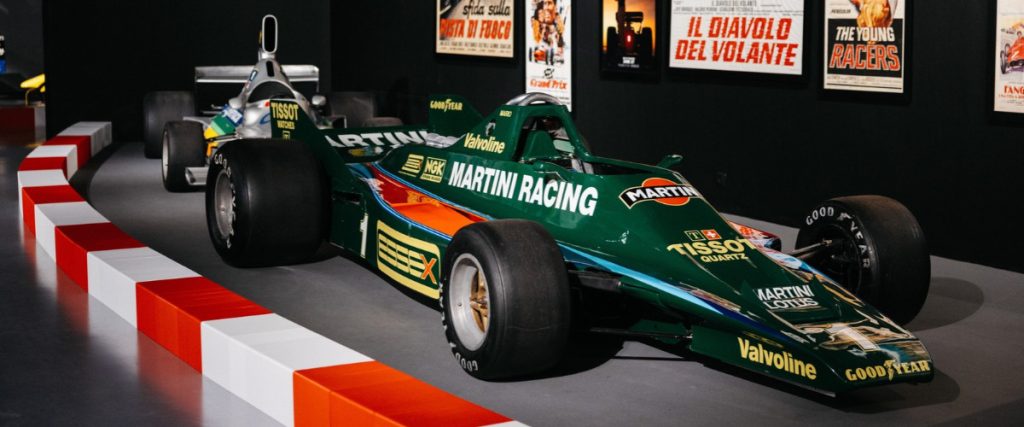 Green Martini Racing Formula 1 car on display in a museum with vintage posters on the wall and red-white barriers in the foreground,